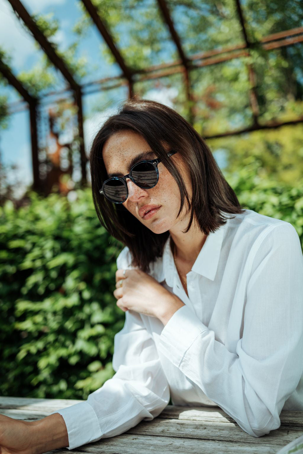 Woman with black sunglasses and white blouse, sitting at a wooden table outdoors, street style